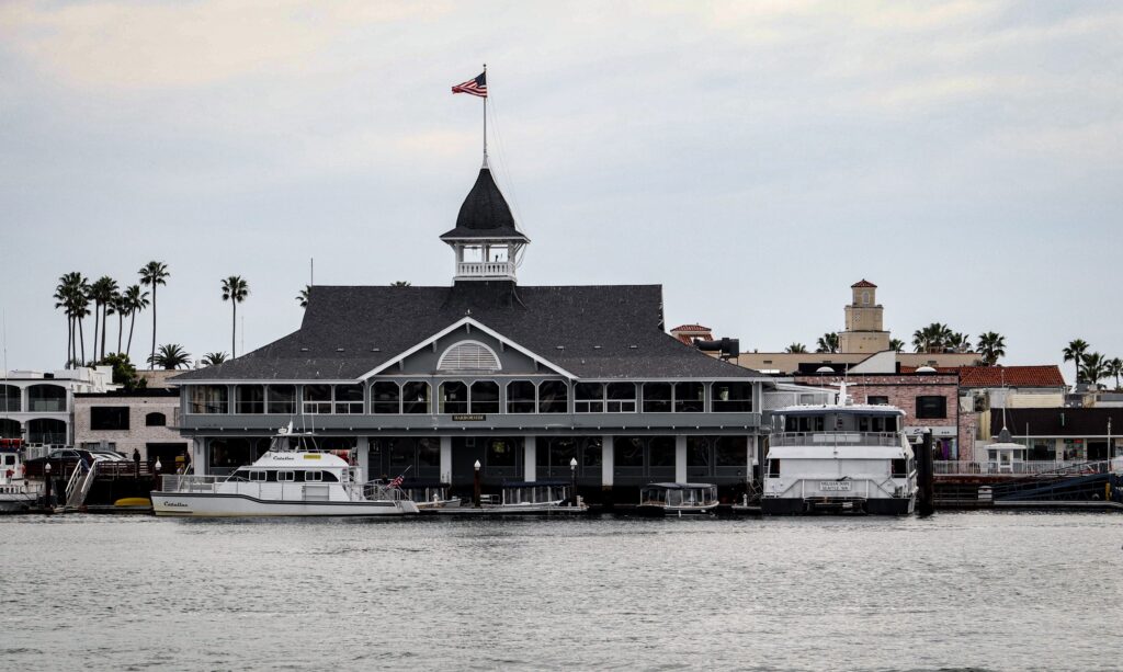 a waterfront view of a grand historic grey building with a Victorian-style facade and a prominent dark cupola spire flying the american flag